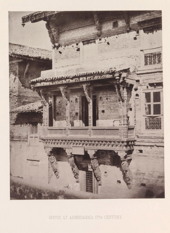 Façade of a house with a balcony in Ahmedabad, Gujarat, India, page, photomechanical print, title: 'HOUSE AT AHMEDABAD, 17TH CENTURY.', number: 'II.', Ahmedabad (city), maker: Heliotype Printing Company, Boston (Massachusetts), c. 1875 - in or before 1885, photographer, North America, Massachusetts, paper, photoaquatint, height 230 mm x width 189 mm, urban housing, balcony, façade (of house or building), A sepia photograph of a brick building facade featuring a carved wooden balcony with ornate columns and decorative brackets, a tiled roof edge with a hanging ornament, rectangular windows with barred grilles, and a textured brick surface with various holes and repairs.