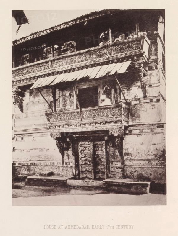 Façade of a house with a balcony in Ahmedabad, Gujarat, India, page, photomechanical print, title: 'HOUSE AT AHMEDABAD, EARLY 17TH CENTURY.', number: 'XXII.', Ahmedabad (city), maker: Heliotype Printing Company, Boston (Massachusetts), c. 1875 - in or before 1885, photographer, North America, Massachusetts, paper, photoaquatint, height 233 mm x width 182 mm, urban housing, balcony, façade (of house or building), A monochrome photograph of an ornate building façade showing a carved wooden balcony with decorative panels and a slanted awning supported by angled beams, an open window or doorway behind the balcony with a blurred light-toned figure or object visible inside, elaborately carved pillars and brackets beneath the balcony, a pair of intricately carved double doors at street level framed by decorative lintels, worn stone steps and a narrow pavement in front, weathered plastered walls with horizontal decorative bands, and an upper storey with a long carved frieze and a projecting ro...