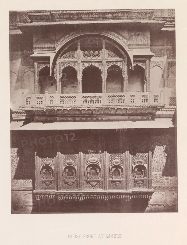 Façade of a house with a balcony in Ajmer, Rajasthan, India, page, photomechanical print, title: 'HOUSE FRONT AT AJMERE.', number: 'VI.', Ajmer, maker: Heliotype Printing Company, Boston (Massachusetts), c. 1875 - in or before 1885, photographer, North America, Massachusetts, paper, photoaquatint, height 231 mm x width 189 mm, urban housing, balcony, façade (of house or building), A sepia-toned photograph of a building facade showing ornate carved stonework and a projecting balcony with patterned balustrades and arched openings. The upper section features a large central arch flanked by smaller arches, each with decorative latticework and scalloped arch details, supported by slender columns with intricate carvings. Below the balcony is a row of elaborately carved panels with recessed arched niches and geometric and floral motifs. The stone surfaces display textured patterns and ornamental friezes, with shadows creating contrast across the architectural details.