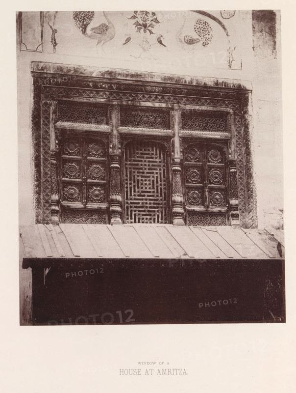 View of a window with ornaments in Amritsar, Punjab, India, page, photomechanical print, title: 'WINDOW OF A, HOUSE AT AMRITZA.', number: 'IX.', Amritsar, maker: Heliotype Printing Company, Boston (Massachusetts), c. 1875 - in or before 1885, photographer, North America, Massachusetts, paper, photoaquatint, height 210 mm x width 179 mm, anthropomorphic ornament, window, A photographic image of an ornate wooden window set into a wall, showing carved panels with floral rosette motifs and geometric latticework, fluted columns and decorative borders framing the window, a painted frieze above with bird and floral designs, a sloped metal awning below with visible seams, and a dark recessed opening beneath the awning.