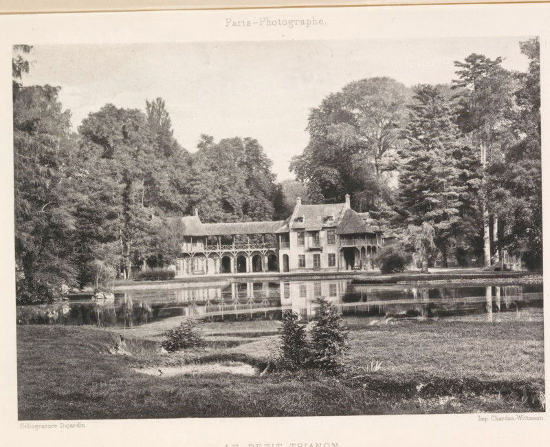 View of the Hameau de la Reine, Le Petit Trianon, page, photomechanical print, photographer: Dujardin, Versailles, maker: Chardon-Wittmann, c. 1881 - in or before 1891, paper, photoaquatint, height 122 mm x width 172 mm, palace, Le Hameau de la Reine, A monochrome image showing a large two-story building with arched openings on the ground level, a covered balcony supported by wooden posts on the upper level, and a steeply pitched roof with chimneys, set at the edge of a reflective body of water; dense trees of varying heights and shapes surround the building on both sides, and the water in front of the building mirrors its facade and the trees, with a grassy foreground containing small shrubs and a gently sloping bank.
