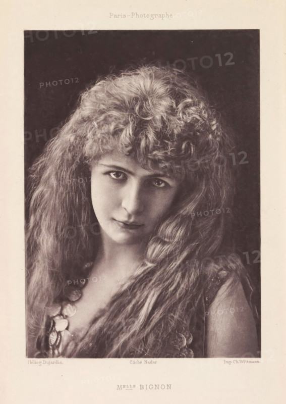 Melle Bignon, Portrait of Louise Bignon, page, photomechanical print, photographer: Félix Nadar, Paris, cliché maker: Dujardin, France, printer (person): Charles Wittmann, c. 1884 - in or before 1894, printer (person), paper, photoaquatint, height 155 mm x width 111 mm, Person, Louise Bignon, portrait of actor, actress, A sepia-toned photographic portrait of a young woman framed by a dark rectangular background, showing her head and upper torso; she has long, wavy hair with a voluminous, curly fringe and soft waves cascading over her shoulders, visible skin tones of a pale complexion, large eyes looking slightly upward, a straight nose, and closed lips with a faint neutral expression; she wears a garment with textured fabric and a necklace made of round, reflective discs that rest on her chest, and the image is bordered by a lighter margin.