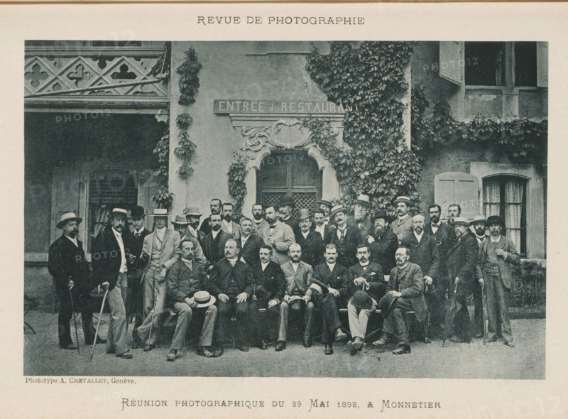 Group portrait of unknown men, Réunion photographique de 29 Mai 1892 a Monnetier, page, photomechanical print, inscription: 'Revue de Photographie', maker: Antoine Elie Chevalley, 1892-05-29, paper, collotype, photoprint, height 115 mm x width 179 mm, anonymous historical persons depicted in a group, in a group portrait, A large group of men posed in front of a building façade covered in climbing ivy and decorative stonework, many wearing suits and hats and some holding walking sticks, with a carved doorway centered behind them and shuttered windows visible to the sides, a patterned balcony railing at the upper left, and a bench along which several seated men are arranged while others stand around them.