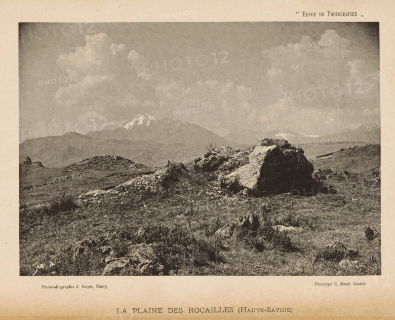 View of a landscape with a mountain range with snow-capped peaks in the background, page, photomechanical print, maker: Jules Royer, maker: E. Mazel, c. 1887 - in or before 1892, paper, collotype, photogravure, height 114 mm x width 163 mm, landscapes, mountains, A sepia-toned landscape photograph showing a rocky foreground with scattered low vegetation and uneven ground, a prominent large boulder and smaller rock formations in the midground, distant rolling hills and a snow-capped mountain peak under a wide sky filled with billowing clouds.