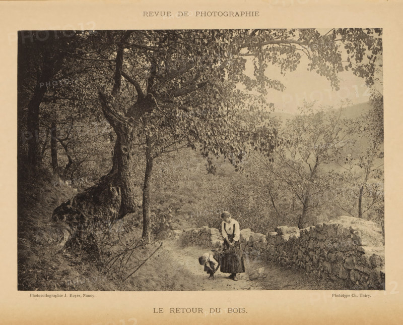 Le redactie du bois, Two unknown young women on a path near a forest, page, photomechanical print, maker: Jules Royer, maker: C. Thiry, c. 1887 - in or before 1892, paper, collotype, photoprint, height 117 mm x width 166 mm, adolescent, young woman, maiden, road, path, trees, A sepia-toned photograph shows a wooded path lined by a low stone wall with uneven rocks, trees with dense leafy branches arching overhead, and a steep embankment on one side; two girls in long dresses stand on the path near the wall, one bending down toward the ground while the other stands upright holding an object and looking down, and scattered foliage and distant hills are visible through the trees.