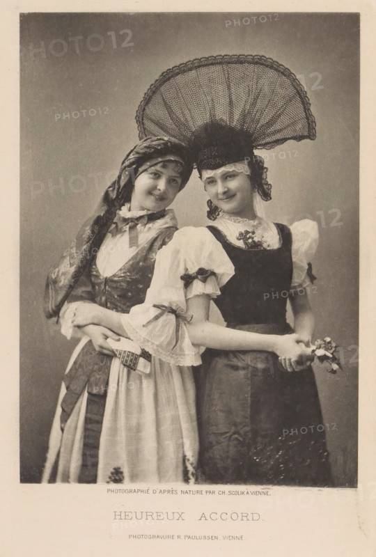 Heureux accord, Portrait of two unknown young women in traditional costumes, page, photomechanical print, photographer: Charles Scolik, maker: Richard Paulussen, c. 1887 - in or before 1892, paper, photoaquatint, height 155 mm x width 110 mm, historical persons, folk costume, regional costume, adolescent, young woman, maiden, Two young women stand close together facing the camera, both wearing traditional-style dresses with puffed sleeves and aprons; the woman on the left wears a patterned headscarf tied around her head and a dress with a patterned bodice and a light apron, while the woman on the right wears a dark bodice over a light blouse and a large ornate lace headdress that fans out above her head; both women have slight smiles and their arms are linked at the waist, with the rightmost woman holding a small cluster of flowers in her hands.