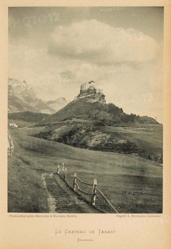 View of a castle in Tarasp, Le Château de Tarasp, page, photomechanical print, photographer: A. Bonnard, maker: Brunner & Hauser, c. 1887 - in or before 1892, paper, collotype, photoprint, height 159 mm x width 119 mm, landscape with tower or castle, (high) hill, A sepia-toned photograph showing a hilltop fortress perched on a rocky outcrop surrounded by rolling grassy slopes and scattered trees, with a winding dirt path and a wooden split-rail fence leading up toward the hill, distant jagged mountains visible on the left, a small building near the base of the hill, and a sky filled with large soft clouds.