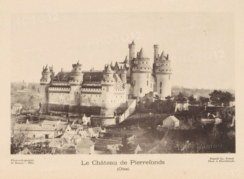 View of the Castle of Pierrefonds, Le Château de Pierrefonds, page, photomechanical print, photographer: Ls. Sorret, maker: H. Besson, c. 1887 - in or before 1892, paper, collotype, photoprint, height 104 mm x width 165 mm, landscape with tower or castle, castle, Pierrefonds, A large stone castle with multiple round towers and conical roofs sits on a raised mound above a cluster of small buildings and trees, showing fortified walls with battlements and rows of windows; steep stair-like pathways and terraces lead up to the castle, while rooftops of nearby houses and patches of open land surround the foreground, and a pale sky fills the upper area of the image.