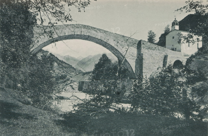 Pont de Neubrücke - Vallée de la Viège, View of the bridge of Neubrück, page, photomechanical print, maker: Frédéric Thévoz, maker: F. Thévoz & Cie., c. 1888 - in or before 1893, paper, collotype, photoprint, height 118 mm x width 174 mm, mountains, bridge, Switzerland, A landscape photograph showing a stone arch bridge spanning a river with vegetation in the foreground and on the riverbanks, leafy branches framing the top of the scene, a steep tree-covered slope to the left, and a white building with a small tower situated near the bridge on the right side, with distant mountain peaks visible through the arch.