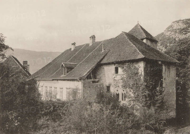 L'ancien prieuré de Talloires, View of a priory in Talloires, page, photomechanical print, maker: Eugène Demole, maker: Brunner & Hauser, c. 1888 - in or before 1893, Eugène Demole, Brunner & Hauser, paper, collotype, photoprint, height 111 mm x width 152 mm, abbey, monastery, convent, Roman Catholic church, Talloires, A sepia-toned photograph shows a large stone building with steep, tiled roofs and several chimneys, partially covered in climbing vegetation; the facade includes shuttered windows and a section with narrow arched windows set in rough stone; dense foliage and bushes fill the foreground and flank the building on the sides; a distant hillside or mountain rises behind the structure under a pale sky.
