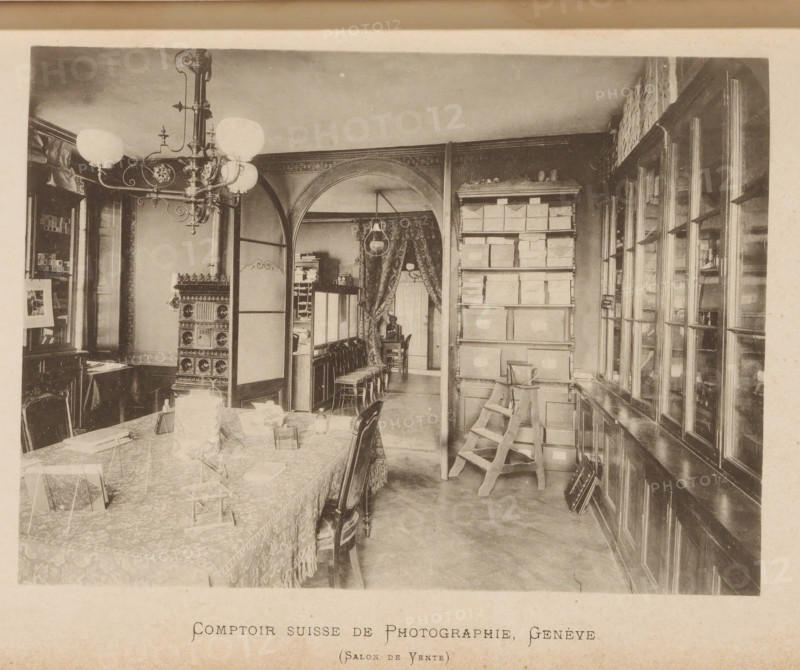 Comptoir suisse de photographie - Salon de Yente, Interior of a Swiss photography shop, page, photomechanical print, maker: F. Thévoz & Cie., c. 1888 - in or before 1893, paper, collotype, photoprint, height 114 mm x width 151 mm, Print on second and third page after page 131., lighting, lamps, edifice, eg: public building, department store - AA - civic architecture: inside, A sepia-toned interior scene shows a room with a long table covered by a patterned cloth and fringed edge, chairs pushed around it, and small display stands and objects arranged on the table. A large ornate chandelier with globe lamps hangs from the ceiling near a decorative stove with ornate panels. An arched doorway divides the space, revealing a curtained area with a hanging lamp and a row of stacked chairs and a small table or desk beyond. To the right, tall shelving holds numerous stacked boxes and a wooden step ladder leaning against them, and a wall of glass-fronted cabinets or windows runs along the far rig...
