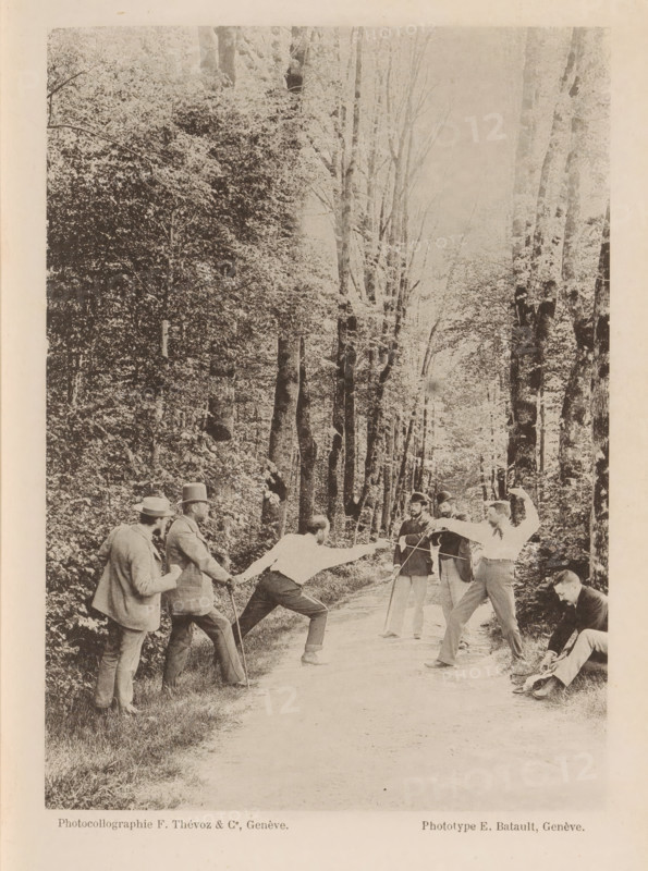 A group of unknown men, two of whom are fencing on a path through the forest, Action, page, photomechanical print, maker: F. Thévoz & Co., maker: E. Batault, c. 1888 - in or before 1893, paper, collotype, photoprint, height 162 mm x width 116 mm, lighting, lamps, fencing (sport), edifice, eg: public building, department store - AA - civic architecture: inside, A sepia-toned photograph shows a tree-lined dirt path with tall trees and dense foliage on both sides, creating a canopy overhead. On the path a group of men are engaged in a fencing duel, two men facing each other in lunging stances with arms extended and foils raised, while several other men stand nearby watching; some wear suits and hats, one holds a cane, and another is seated at the path edge adjusting something near his feet. The scene captures varied postures and focused expressions among the participants against the backdrop of the wooded avenue.