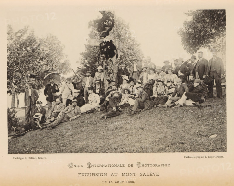 Union Internationale de Photographie - Excursion au Mont Salève, Group portrait of members of the Union Internationale de Photographie, page, photomechanical print, maker: Jules Royer, maker: E. Batault, 1893-08-20, paper, collotype, photoprint, height 105 mm x width 160 mm, anonymous historical persons depicted in a group, in a group-portrait, Salève, Mont, A sepia-toned photograph shows a large group of men and women gathered outdoors on a grassy slope in front of a large tree. The people are arranged sitting, reclining, and standing, many wearing period clothing including suits, hats, and long dresses; several women wear wide-brimmed hats and some men wear bowler or brimmed hats. A few people hold umbrellas, canes, or walking sticks, and one person appears to be seated on a bench at the right edge. Trees and foliage form the background, and the ground in the foreground is grassy with scattered leaves. The image has a wide white margin around it.