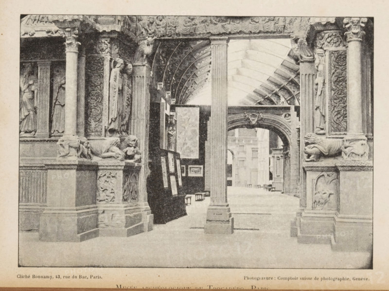 Musée archeologique du trocadéro, Paris, View of an entrance to a room of the Musée archeologique du trocadéro, page, photomechanical print, workshop stamp: 'No 7887, post, tenebras, spero, lucem', cliché maker: Bonnamy, maker: Comptoir Suisse de Photography, c. 1889 - in or before 1894, Comptoir Suisse de Photographie, paper, autotype, height 123 mm x width 173 mm, column, pillar, architecture, exhibition, art, A sepia-toned indoor scene shows a vaulted gallery with ornate stone columns and carved architectural details. Sculpted figures are set into niches and decorative relief panels run vertically along the columns. Stylized animal sculptures rest on ledges near the column bases. An archway with carved ornamentation frames a view into a distant interior space with additional columns and architectural elements. Framed artworks are displayed on easels and leaning against a dark partition beneath the vaulted ceiling. The floor appears smooth and open beneath the sequence of arches and ...