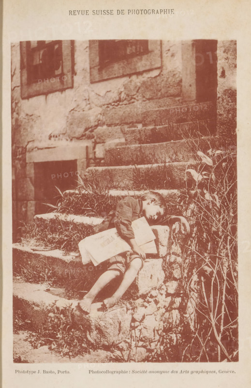 Noite de Natal, Unknown boy lying on a staircase without shoes, page, photomechanical print, maker: Joaquim Basto, maker: Société Anonyme des Arts Graphiques, c. 1889 - in or before 1894, paper, collotype, photoprint, height 164 mm x width 115 mm, boy (child between toddler and youth), flight of steps, A sepia-toned photograph shows a young boy reclining on worn stone steps beside a rough plaster wall and a dark doorway, his head resting on one arm and his other arm draped over the edge of a step, a folded newspaper held against his chest, his bare feet and legs extending down the steps amid patches of grass and small plants, framed by a vertical cluster of leafy stems on the right and weathered window openings higher on the wall.