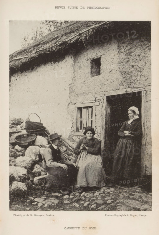 Two unknown women and an unknown man in front of a building, Causette du soir, page, photomechanical print, maker: M. Bovagne, maker: Jules Royer, c. 1890 - in or before 1895, paper, collotype, photoprint, height 163 mm x width 122 mm, adult woman, adult man, open door, A black and white photograph shows part of a rustic building with a thatched roof and a textured wall, a small window with wooden framing, and a doorway with a wooden lintel. In front of the building are three people: a seated man wearing a hat and vest, a seated older woman in a dress and apron holding her hands together, and a standing woman in a long dress and headscarf leaning in the doorway with her arms crossed. To the left of the seated man there are woven baskets placed on a pile of stones and some scattered rocks and cobbles on the ground in front of the group.