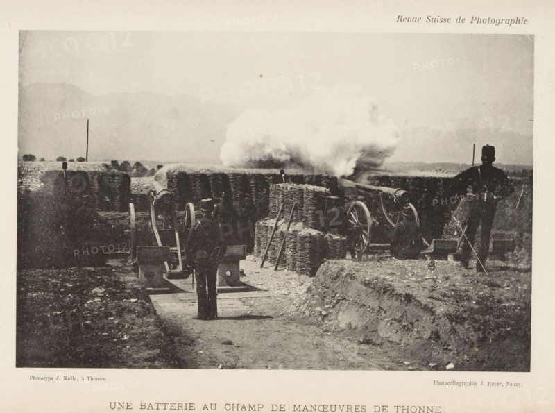 Une Batterie au Champ de Maneuvers de Thonne, Two gunners at Thonne, France, page, photomechanical print, maker: J. Kolla, maker: Jules Royer, c. 1891 - in or before 1896, paper, collotype, photoprint, height 113 mm x width 173 mm, firearms: cannon, Thonne, A grainy black and white scene showing a dirt path flanked by low earthworks and scattered rocks leading toward a line of artillery pieces behind woven wicker or gabion barriers, with plumes of smoke billowing from one of the guns; several uniformed figures stand or kneel near the guns, one figure facing the camera on the path and another standing to the right with a long object in hand; wooden wheels and gun carriages are visible among stacked wicker bundles and upright poles, and distant low hills or mountains form a pale backdrop under a wide sky.