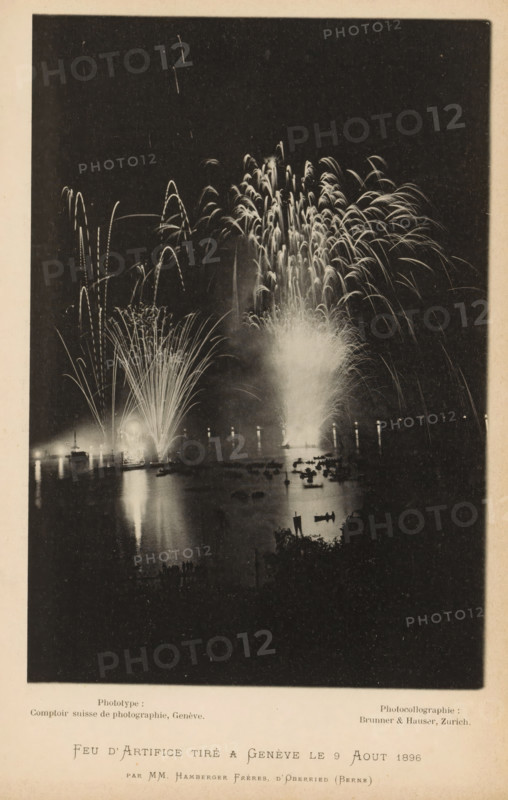 Feu d'Artifice tiré a Genève le 9 Aout 1896, View of fireworks over the harbor of Geneva, page, photomechanical print, inscription: 'par MM. Hamberger Frères, d'Oberried (Berne)', photographer: Hamberger Frères, Genève, maker: Comptoir Suisse de Photographie, maker: Brunner & Hauser, Zürich (city), 1896-08-09, paper, collotype, photoprint, height 180 mm x width 123 mm, harbour, bonfire, fire-works - AA - festivities on the water, Geneva, A nighttime scene showing fireworks exploding over a body of water, with bright cascades and arcing trails of light rising and reflecting on the water surface; several boats and small silhouettes are visible on the water, and a shoreline with darker shapes and vegetation appears in the lower foreground; the image has a dark surrounding border and lighter margins around the edges.