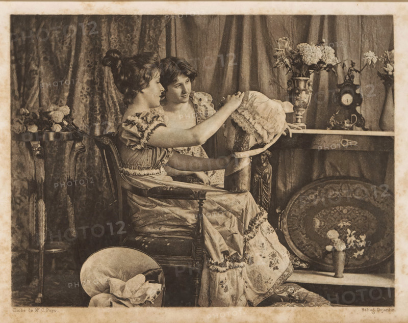 Two women examining a hat, page, photomechanical print, cliché maker: Constant Puyo, printer (person): Dujardin, c. 1891 - in or before 1896, photographer, printer (person), paper, photoaquatint, height 125 mm x width 164 mm, adult woman, head-gear: hat, Two young women sit closely on an upholstered armchair in front of patterned drapery, both wearing ruffled dresses; one woman faces sideways while the other looks toward her, and they hold a large bonnet with lace and ribbon between them. To the right a mantel or table displays a tall ornate vase filled with flowers, a decorative clock, and another floral arrangement in a narrow vase, while a round decorative tray and a small urn with flowers sit beneath the table. To the left a small side table holds a bouquet of flowers, and a hat with fabric sits on the floor near the chair. The overall image has a sepia tone and shows detailed textures of fabric, wood carving on the chair, and floral patterns.