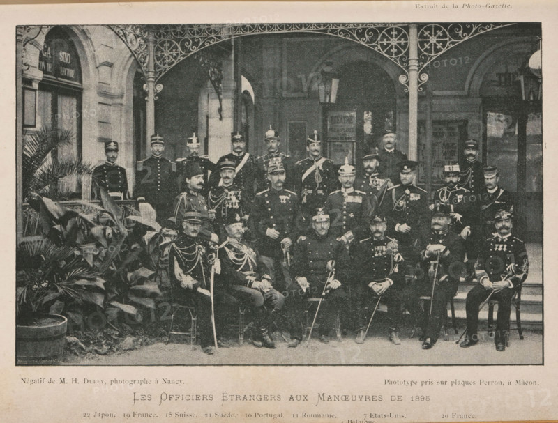 Group portrait of foreign officers, Les Officiers Étrangers aux Maneuvers de 1895, page, photomechanical print, inscription: '22 Japon. 19 France. 15 Suisse. 21 Suede. 10 Portugal. 11 Roumania. 7 Etats Unis. 20 France., 6 T... 4 Belgique', inscription: 'Extrait de la Photo-Gazette.', inscription: 'Phototype pris sur plaques Perron, à Màcon.', photographer: Henry Dufey, cliché maker: anonymous, 1895, paper, autotypie, height 115 mm x width 179 mm, Print on third unnumbered page after page 80., anonymous historical persons depicted in a group, in a group-portrait, A sepia-toned photograph shows a large group of uniformed men posed on steps and chairs beneath an ornate metal awning in front of a stone building with arched doorways and windows; many men wear peaked caps or helmets and military jackets adorned with medals, sashes, and braided cords, and several hold swords or canes; potted plants with broad leaves occupy the foreground at one side, and decorative ironwork frames the top of ...