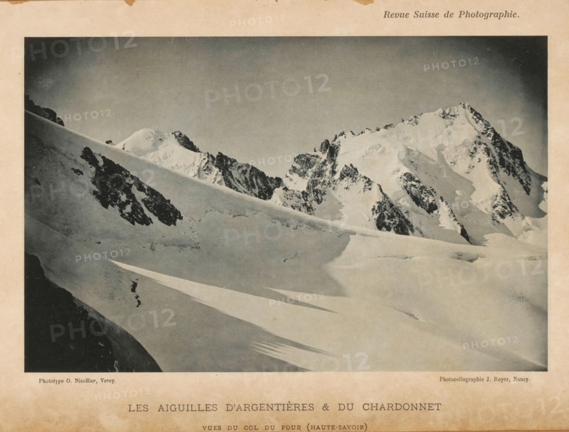 View of the Aiguille d'Argentières and the Aiguille de Chardonnet in the French Alps, Les Aiguilles d'Argentières & du Chardonnet, page, photomechanical print, inscription: 'vues du col du four (Haute-Savoie)', maker: O. Nicollier, maker: Jules Royer, c. 1891 - in or before 1896, paper, collotype, photoprint, height 111 mm x width 172 mm, mountain range, snow peaks, Argentière glacier, Aiguille du Chardonnet, A black and white photograph of a snowy mountain scene framed by a pale border, showing a sweeping snowfield in the foreground with long diagonal shadows, a steep snow-covered slope cutting across the midground, and jagged rocky peaks capped with snow rising in the background under a clear sky.