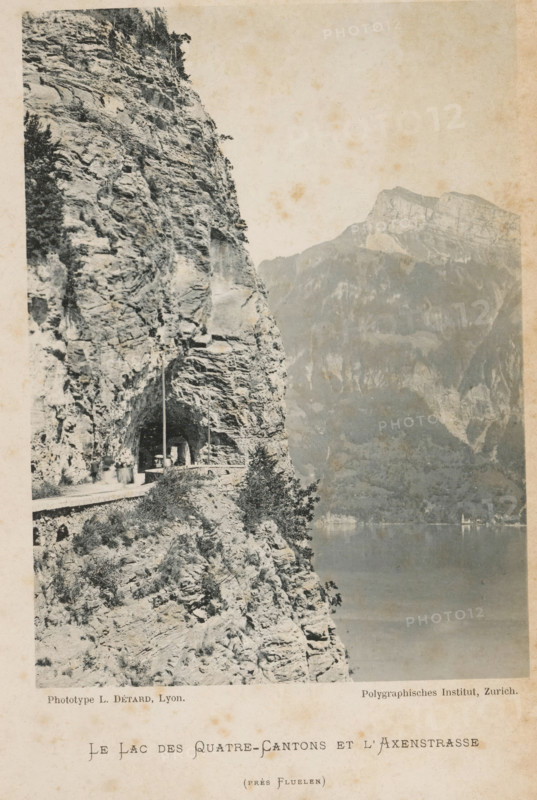 Le Lac des Quatre-Cantons et l'Axenstrasse, View of the Axenstrasse and Lake Lucerne, page, photomechanical print, maker: L. Détard, maker: Polygraphisches Institut AG, c. 1892 - in or before 1897, L. Détard, Polygraphisches Institut AG, paper, collotype, photoprint, height 163 mm x width 119 mm, lake, mountains, Lake Lucerne, Axenstrasse, A monochrome photograph shows a steep rocky cliff face with rugged strata and sparse vegetation along a narrow road carved into the rock, the road passing through a short tunnel in the cliff where a lamp post and a small figure are visible; below the cliff edge the terrain drops toward a calm lake whose surface reflects muted tones, and opposite the lake a distant mountainside with layered rock faces rises under a pale sky.