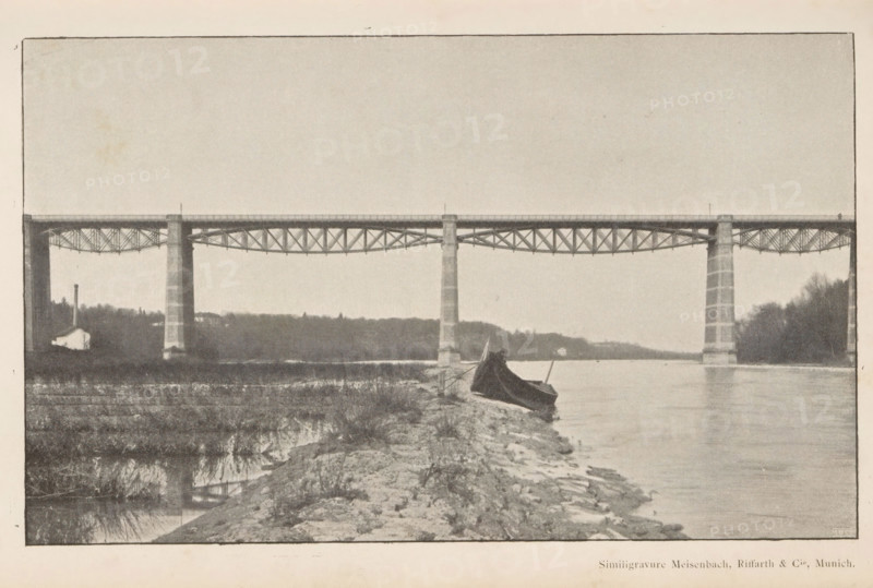 Pont sur l'Isar, View of a bridge over the river Isar, page, photomechanical print, Isar, cliché maker: Meisenbach, Riffarth & Cie., Munich, c. 1892 - in or before 1897, Meisenbach, photographer, cliché maker, Bavaria, paper, autotypie, height 115 mm x width 189 mm, river, bridge, A wide river scene with a long metal bridge spanning across the water supported by tall stone piers, a shoreline in the foreground with rocky ground and sparse grass, a small boat pulled up on the shore partially covered by a dark tarp, distant tree-covered banks on both sides of the river, and a calm water surface reflecting parts of the bridge and surrounding landscape.