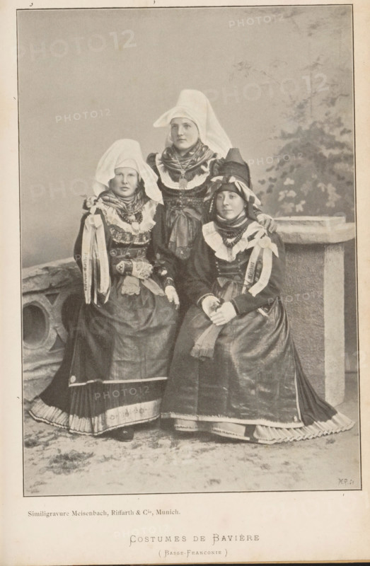 Costumes de Bavière (Basse-Franconie), Portrait of three unknown women in Bavarian costumes, page, photomechanical print, cliché maker: Meisenbach, Riffarth & Cie., Munich, c. 1892 - in or before 1897, photographer, Bavaria, paper, autotypie, height 180 mm x width 125 mm, folk costume, regional costume, anonymous historical persons depicted in a group, in a group-portrait - BB - woman, Three women are posed together wearing long dresses with full skirts and layered aprons, high collars, and ornate necklaces; each has a head covering with one tall woman standing behind two seated women, the standing woman wearing a pointed white headdress that drapes down behind her and a layered scarf around her neck, the seated women wearing different shaped headpieces with ribbons and decorative bands, one seated woman resting her hand on the other's shoulder while the other clasps her hands in her lap; the dresses show patterned trims and pleated underskirts at the hems, and the backdrop includes a ...