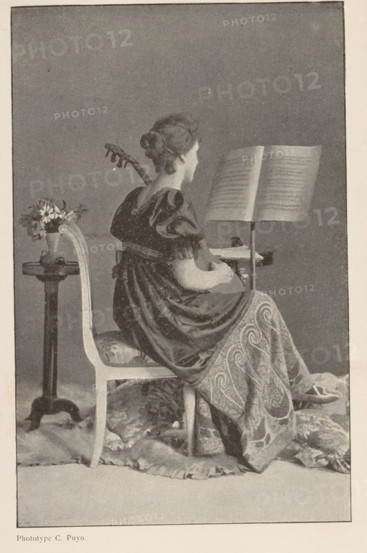 La Musique, Portrait of an unknown woman playing the guitar, page, photomechanical print, inscription: '(Extrait de Photo-Gazette.)', maker: Constant Puyo, c. 1892 - in or before 1897, paper, autotypie, height 169 mm x width 108 mm, historical persons not known by name - BB - woman, cithern, mandolin, guitar, balalaika (artist at work), A seated woman is shown from behind and in profile, wearing a dark, short-sleeved dress with a patterned wrap draped over her lower body; her hair is styled in an updo and a stringed instrument rests against her back with her hands positioned near its body. She sits on a carved chair with an upholstered seat and a floral cushion at her feet, a music stand in front of her holding open sheets, and a small pedestal table to her side holds a vase filled with flowers. The floor beneath her is covered with draped fabric and cushions, and the scene is framed by a plain backdrop.