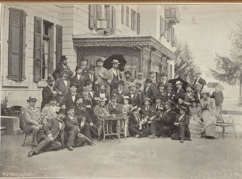 Réunion aux Avants, sur Montreux, le 17 Mai 1899, Group portrait of a reunion near Montreux, Switzerland, probably from the Sociéte des Photographes Suisses, page, photomechanical print, maker: Edouard Potterat, maker: Brooke & Kühn, 1899-05-17, paper, autotypie, height 125 mm x width 170 mm, anonymous historical persons depicted in a group, in a group-portrait, Montreux, A sepia-toned photograph shows a large group of people arranged in front of a building with shuttered windows and a decorative awning. Most individuals wear suits and hats, with some women in dresses and hats seated among them. Several men hold canes or umbrellas, one umbrella is open above the group, and a few people are seated at a small round table with ornate metalwork. The building facade features multiple tall windows with shutters and a covered porch area with decorative trim. In the background there are trees and a light-colored open area, and a few chairs and benches are visible at the edges of the scene.