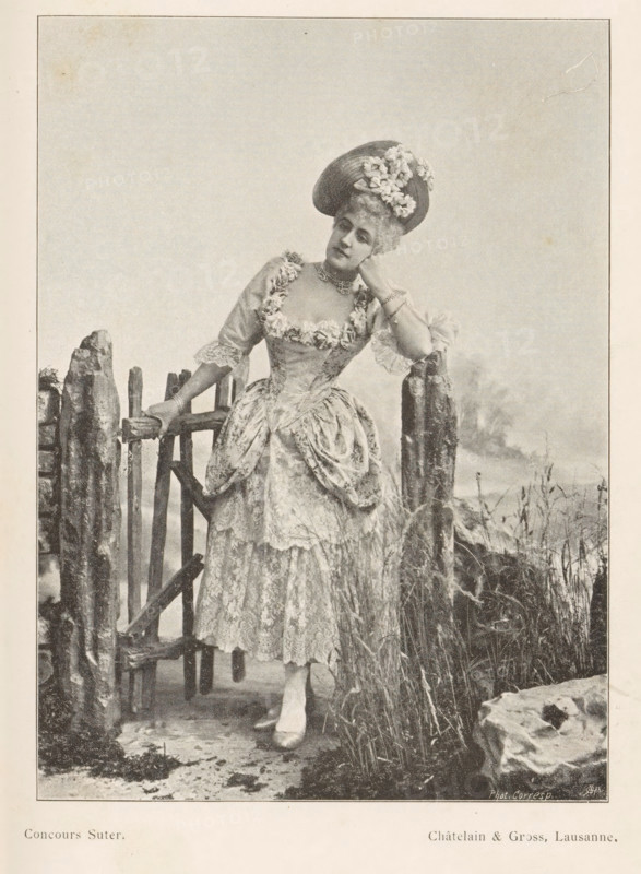 Portrait of an unknown woman at a fence, page, photomechanical print, inscription: 'Phot. Corresp.', inscription: 'Concours Suter.', monogram: 'AG ph.', maker: Châtelain & Gross, cliché maker: unknown, c. 1894 - in or before 1899, paper, autotypie, height 143 mm x width 104 mm, historical persons not known by name - BB - woman, gate, entrance, A sepia-toned photograph of a woman standing by a rustic wooden gate and fence, wearing an ornate dress with lace and floral trim and a wide skirt with gathered overskirt, a hat decorated with flowers, gloves, a choker necklace, and a bracelet; she leans with one elbow on a fence post and rests her hand against her cheek while the other hand holds the gate, grasses and rough stones surround the fence, and the background is a plain sky.
