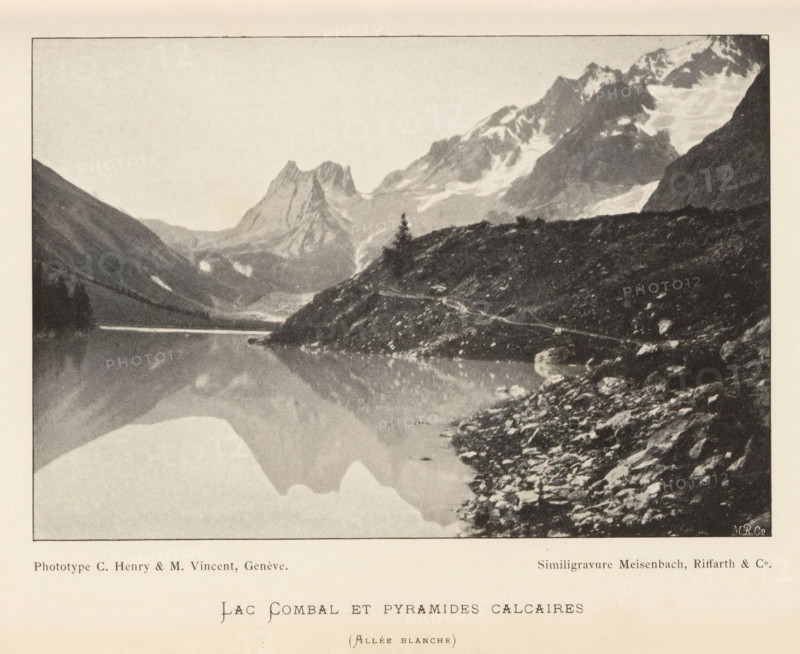 View of a mountain lake, probably in Switzerland, Lac Combal et pyramides calcaires, page, photomechanical print, inscription: '(Allée blanche)', maker: Henry, C. & Vincent, M., cliché maker: Meisenbach, Riffarth & Cie., Munich, c. 1894 - in or before 1899, Bavaria, paper, autotypie, height 101 mm x width 149 mm, lake, mountains, A sepia-toned landscape photograph showing a calm lake with a clear reflection of surrounding mountains and sky, steep rocky shores in the foreground with scattered stones and a narrow path running along the water's edge, a lone conifer tree on a protruding ridge, layered mountain peaks with patches of snow and glaciers in the background, and a pale, lightly textured sky above.