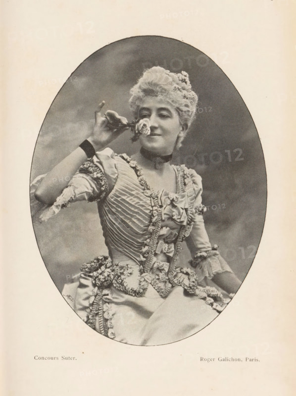 Portrait of an unknown woman, page, photomechanical print, monogram: 'AG ph.', inscription: 'Phot. Corresp.', inscription: 'Concours Suter', maker: Roger Galichon, cliché maker: unknown, c. 1894 - in or before 1899, paper, autotypie, height 136 mm x width 104 mm, historical persons not known by name - BB - woman, cut flowers, nosegay, bunch of flowers, A sepia-toned oval photograph shows a woman in a decorative, ruffled dress with layered fabric and floral trim, seated and holding a small flower near her face with one hand; she has styled, curly hair adorned with small flowers, a choker around her neck, and a faint smile, while the background within the oval is plain and softly shaded.