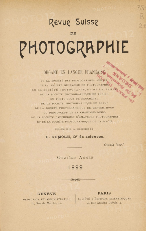 Revue suisse de photographie, Société genevoise de photographie, et al., book, collector's mark: 'Dr. Frans de Vos, 54, Ter Rivierenlaan, DEURNE, Telst.: 5.6.90 - Postcheckr. .04.1..', annotation: 'NOV, 1994', annotation: 'appareils à main. Page 45.', name: 'Jer. Van Severen, Ruysselede', editor (person who revises): Eugène Demole, 1899, Eugène Demole, editor (person who revises), textile materials, pasteboard, biological material, paper, linen (material), leather, printing, shell marbled paper, agate marbled paper, lithography, autotype, collotype, height 234 mm x width 168 mm x thickness 27 mm, The image shows an aged, beige paper cover with a large stylized title near the top and multiple lines of printed text arranged vertically down the center; there is a diagonal stamped mark in a darker color on the right side of the page and a small decorative flourish near the middle; the paper has a slightly textured, worn appearance and even margins around the printed content.