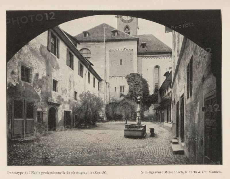 Fraumünsteramt, Courtyard of the Fraumünster Monastery in Zurich, page, photomechanical print, maker: l'Ecole professionelle de photographie, cliché maker: Meisenbach, Riffarth & Cie., Munich, c. 1895 - in or before 1900, l'Ecole professionelle de photographie, Meisenbach, cliché maker, Bavaria, paper, autotypie, height 109 mm x width 152 mm, courtyard, cloisters, monastery, Zürich (city), A cobblestone courtyard framed by a dark arched opening shows aged buildings with textured, weathered facades and shuttered windows lining both sides, a central stone fountain with a tall ornate column and rectangular basin sits in the open space, a large building with a steep roof and small dormer windows rises at the far end behind a leafy tree, narrow doorways and small steps lead into the buildings, and the scene is rendered in a monochrome, grainy photographic style.