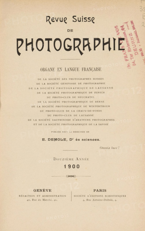 Revue suisse de photographie, Société genevoise de photographie ... et al., book, date: 'Nov 1994', annotation: '... van deveren ...', number: '288', collector's mark: 'Dr. Frans de vos, 64 tot Rivierenlaan, 64, Deurne, ...', editor (person who revises): Eugène Demole, 1900, Eugène Demole, editor (person who revises), textile materials, paper, linen (material), transparent paper, printing, autotype, collotype, height 234 mm x width 172 mm x thickness 29 mm, The image shows a vertical page with a cream background and printed elements arranged from top to bottom, including a large bold decorative word near the top, a horizontal thin line, several lines of smaller uppercase text forming centered blocks, a stamped diagonal mark in a darker color on the right side, a small ornamental divider, and two separated blocks of text near the lower portion with a vertical dividing line between them, all set against a plain background with even margins.