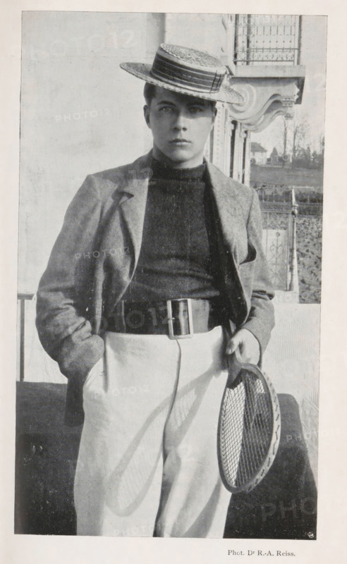 Portrait of an unknown man with a tennis racket, page, photomechanical print, monogram: 'C. Cie', photographer: Rodolphe Archibald Reiss, cliché maker: Corbaz & Cie, c. 1897 - in or before 1902, paper, autotypie, height 178 mm x width 103 mm, historical persons, tennis, lawn-tennis, A black-and-white portrait of a young person standing outdoors wearing a straw boater hat tilted at an angle, a dark high-neck sweater, a light jacket draped over the shoulders, light trousers held by a wide belt with a rectangular buckle, and one hand in a pocket while the other holds a round tennis racket with visible strings; an ornate building facade and a railing appear in the background.