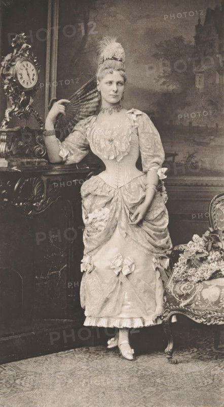 Portrait of an unknown woman, page, photomechanical print, photographer: Fritz Luckhardt, Vienna (possibly), cliché maker: Richard Paulussen, c. 1887 - in or before 1892-02-20, Lower Austria, paper, photoaquatint, height 196 mm x width 109 mm, historical persons not known by name - BB - woman, dress, gown, A full-length portrait of a woman standing beside a carved mantel and an upholstered chair, wearing an ornate gown with a fitted bodice, puffed sleeves, layered skirt with bows and ruffles, and decorative floral accents; she holds a folded fan near her shoulder and wears a feathered headpiece, a choker necklace, earrings, and bracelets, with her hair styled high; the mantel features an elaborate clock and ornamental carving, and the chair has patterned upholstery with a bouquet of flowers resting on it; the background shows a painted scenic backdrop and the floor is covered by a patterned rug.