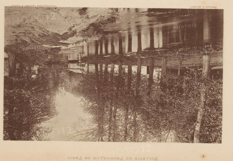 Les Bords de l'Epte, a Gisors, Bank of the river Epte near Gisors, France, page, photomechanical print, inscription: 'Bulletin du Photo-Club de Paris', maker: Paul Bourgeois, maker: Laussedat & Sabatier, c. 1888 - in or before 1893, Paul Bourgeois, Laussedat & Sabatier, paper, collotype, photoprint, height 110 mm x width 180 mm, trees, river, Gisors, An inverted sepia-toned photograph of a tree-lined scene with a body of water reflecting tall trunks and branches, showing dense foliage along the banks, a wooden fence or railing visible near the water, and a building partially seen through the trees.