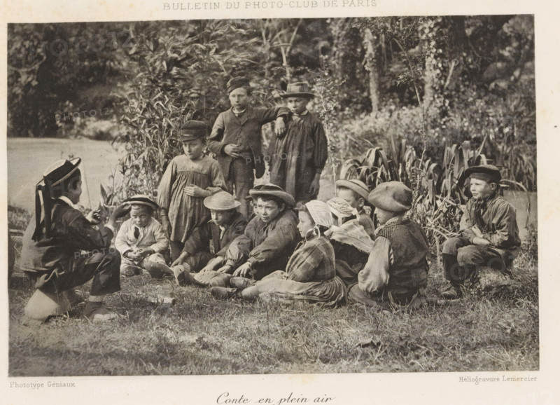Conte en plein air, Boy telling a story to a group of children on a field, page, photomechanical print, inscription: 'Bulletin du Photo-Club de Paris', maker: Paul Géniaux, cliché maker: Lemercier, c. 1888 - in or before 1893, paper, collotype, photoprint, height 113 mm x width 169 mm, park, child between toddler and youth (of undetermined sex), story teller, listening to story teller, A sepia-toned photograph shows a group of children outdoors seated and standing on grass near tall plants and trees, many wearing hats and simple clothing; some children sit cross-legged while others kneel or stand, a few wear bonnets or caps and one child in the foreground gestures with their hands toward the seated group, while the background features dense foliage and a body of water partially visible at the left edge.