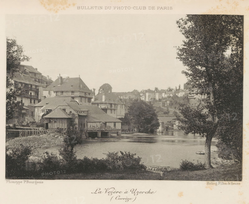 La Vezère à Uzerche, View of the river Vézère in Uzerche, France, page, photomechanical print, inscription: 'Bulletin du Photo-Club de Paris', photographer: Paul Bourgeois, cliché maker: Fillon & Heuse, Paris, c. 1890 - in or before 1895, paper, photoaquatint, height 119 mm x width 170 mm, prospect of village, silhouette of village, river, Uzerche, Vézère, A sepia-toned riverside scene showing clustered riverside buildings with pitched roofs and wooden balconies along the water's edge, a stone-supported structure extending over the river, a calm stretch of water reflecting the scene, a tree-lined riverbank with a large tree in the right foreground, scattered shrubs and vegetation along the near bank, distant houses and hillside structures visible beyond the main buildings, and a stone bridge partially visible spanning the river in the midground.