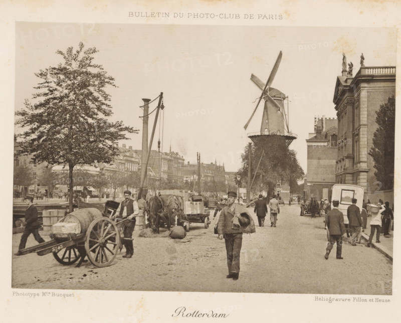 Rotterdam, View of a street in Rotterdam, with a mill in the background, page, photomechanical print, inscription: 'Bulletin du Photo-Club de Paris', photographer: Maurice Bucquet, cliché maker: Fillon & Heuse, Paris, c. 1890 - in or before 1895, paper, photoaquatint, height 120 mm x width 170 mm, street, windmill, A street scene showing a cobblestone road with people walking and standing, some carrying items and one man walking toward the camera holding a bundled object over his shoulder, horse-drawn carts and wooden wagons loaded with barrels and crates, a large tree on the left, a windmill with visible sails near the center-right, a tall building with classical architectural details and statues on its roof on the right, lamp posts and poles with hanging equipment, rows of multi-story buildings in the background, and various street activity with figures in period clothing.