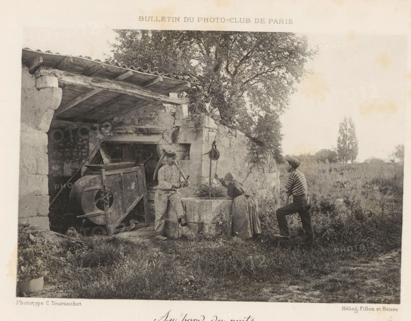 At the well, Two men and a woman around a well, page, photomechanical print, inscription: 'Bulletin du Photo-Club de Paris', maker: C. Touranchet, cliché maker: Fillon & Heuse, Paris, printer (person): Charles Wittmann, c. 1890 - in or before 1895, printer (person), paper, autotype, height 120 mm x width 170 mm, adult woman, adult man, well, A grayscale photograph shows an outdoor scene with a stone structure on the left featuring a tiled overhang and wooden beams, and a large leafy tree rising behind it; in front of the structure there is an old mechanical device with a large wheel and a trough or basin set into a low stone wall; three people stand by the basin, one wearing a cap and light clothing leaning against the wall holding a curved implement, another bending over the basin with a skirt and headscarf, and a third in a striped top and cap standing on the grass with one foot on a low step holding a pole; the foreground is grassy with some low vegetation and a potted plant at the ...