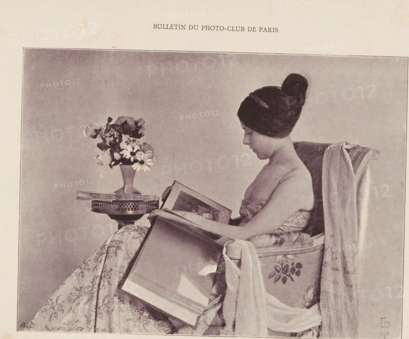 Liseuse, Portrait of an unknown woman reading, page, photomechanical print, monogram: 'AG ph.', maker: Constant Puyo, cliché maker: unknown, printer (person): Draeger & Lesieur, Paris, c. 1890 - in or before 1895, printer (person), paper, autotypie, height 119 mm x width 162 mm, historical persons not known by name - BB - woman, reading, A sepia-toned photograph of a seated woman in profile, wearing her hair in a high bun and a strapless patterned dress, holding and looking at an open large book on her lap; she sits in an upholstered armchair draped with a long fabric, and beside her is a small round table with a vase of mixed flowers on top.
