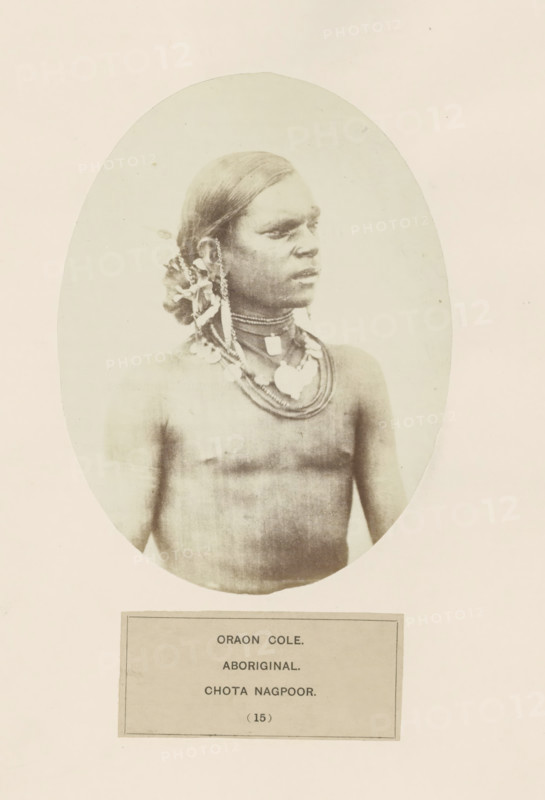 Portrait of an unknown man from the Kurukh community of Chota Nagpur in eastern India, photograph, page, The Kurukh community is also known as Oraon., number: '(15)', inscription: 'ORAON COLE., ABORIGINAL., CHOTA NAGPOOR.', photographer: Benjamin Simpson (possibly), Chota Nagpur, c. 1858 - in or before 1868, Jharkhand, paper, albumen print, height 159 mm x width 120 mm, human races, peoples, nationalities, An oval-framed sepia portrait of a bare-chested person with smooth, dark hair pulled back and adorned with decorative elements at the side, wearing multiple layered necklaces and pendants, looking to the right with a neutral expression against a plain light background, with a rectangular label positioned below the portrait.