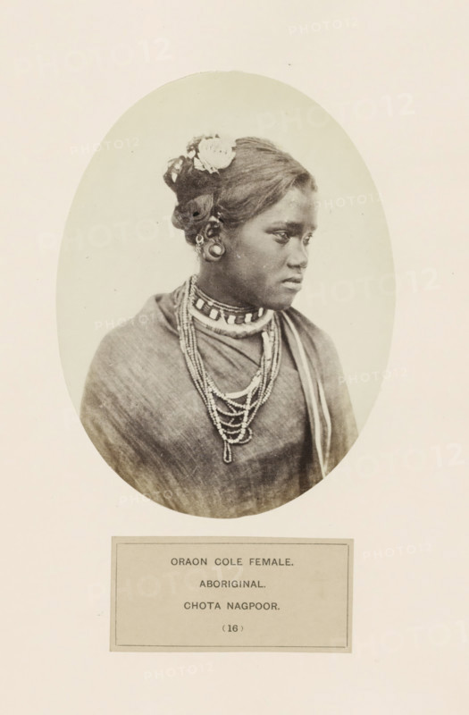 Portrait of an unknown woman from the Kurukh community of Chota Nagpur in eastern India, photograph, page, The Kurukh community is also known as Oraon., inscription: 'ORAON COLE FEMALE., ABORIGINAL., CHOTA NAGPOOR.', number: '(16)', photographer: Benjamin Simpson (possibly), Chota Nagpur, c. 1858 - in or before 1868, Jharkhand, paper, albumen print, height 159 mm x width 120 mm, human races, peoples, nationalities, A sepia-toned oval portrait of a young woman shown from the chest up, turned slightly to her left with a neutral expression, wearing a draped garment and multiple layered beaded necklaces, her hair styled up with a flower tucked near the top and decorative ear ornaments visible, set against a plain light background and mounted on a larger sheet of paper.