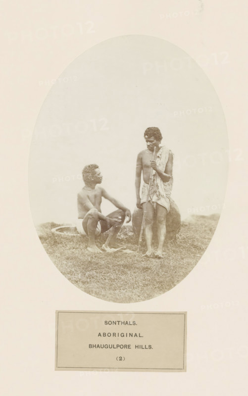 Two unknown men from the Santhal tribe in eastern India, photograph, page, inscription: 'SONTHALS., ABORIGINAL., BHAUGULPORE HILLS.', number: '(2)', Bhagalpur, c. 1858 - in or before 1868, photographer, North India, Bihar, paper, albumen print, height 175 mm x width 130 mm, human races, peoples, nationalities, Jharkhand, Rajmahal hills, An oval-framed sepia photograph shows two barefoot men on grassy ground, one man seated with knees bent and arms resting on them, wearing a short cloth around his waist and looking up toward the standing man, while the standing man wears a draped cloth over one shoulder and around his waist, holds a long wooden staff vertically, and stands beside a large rounded earthen vessel partially visible behind him; the background inside the oval is plain and light, and the image is mounted on a pale rectangular sheet.