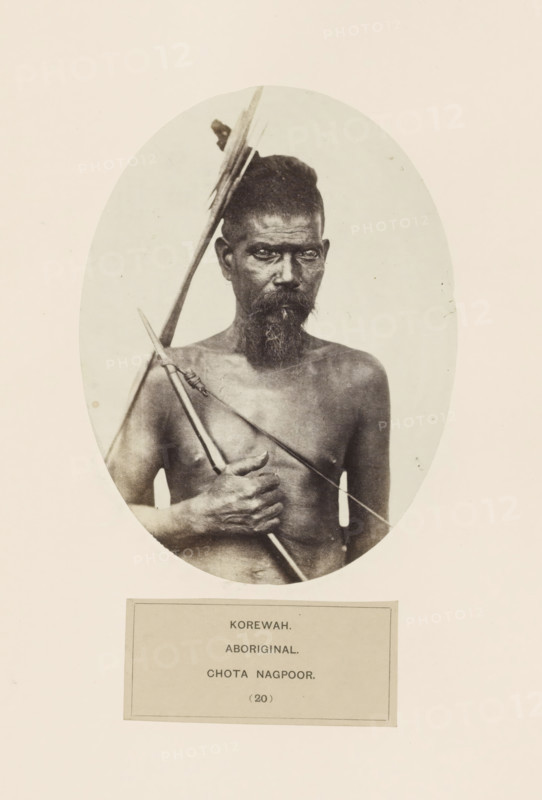 Portait of an unknown man with a bow and arrow from the Korwa tribe of Chota Nagpur in eastern India, photograph, page, number: '(20)', inscription: 'KOREWAH., ABORIGINAL., CHOTA NAGPOOR.', photographer: Benjamin Simpson, Chota Nagpur, c. 1858 - in or before 1868, Jharkhand, paper, albumen print, height 160 mm x width 121 mm, archer's weapons: bow and arrow, human races, peoples, nationalities, An oval black-and-white portrait of a bare-chested man with a narrow face and a beard, his hair tied up, holding a long spear diagonally across his chest and a shorter pointed implement in his hand, with a neutral light background and a rectangular label area below the oval.