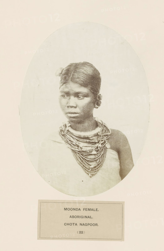 Portrait of an unknown woman from the Munda tribe of Chota Nagpur in eastern India, photograph, page, number: '(22)', inscription: 'MOONDA FEMALE., ABORIGINAL, CHOTA NAGPOOR.', photographer: Benjamin Simpson (possibly), Chota Nagpur, c. 1858 - in or before 1868, Jharkhand, paper, albumen print, height 175 mm x width 130 mm, human races, peoples, nationalities, A sepia-toned oval portrait of a young woman shown from the shoulders up, with dark, smooth hair parted in the middle and pulled back, a small hair ornament visible near one ear, a composed facial expression with eyes looking slightly to one side, layered beaded necklaces draped around her neck and upper chest, and a simple garment covering one shoulder; the portrait is set against a light background with a rectangular label area below.