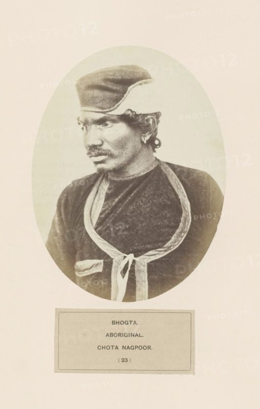 Portrait of an unknown man from the Bhogta community of Chota Nagpur in eastern India, photograph, page, number: '(23)', inscription: 'BHOGTA., ABORIGINAL., CHOTA NAGPOOR.', photographer: Benjamin Simpson (possibly), Chota Nagpur, c. 1858 - in or before 1868, Jharkhand, paper, albumen print, height 160 mm x width 120 mm, human races, peoples, nationalities, head-gear: cap, Oval-framed sepia portrait of a man shown from the chest upward, wearing a rounded cap and a dark garment with a lighter decorative trim forming a looped shape and tied at the front, the man looking slightly to his left with a mustache and short curly hair visible at the ear, the image set against a pale background with a rectangular panel below the portrait.